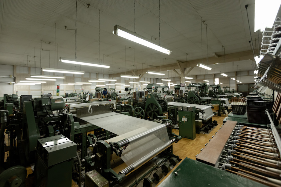 自働織機が並ぶ工場内。部屋いっぱいに、ズラリと並ぶ織り機は60台ほど。数もさることながら、工場内に入った瞬間にその大きな音に圧倒される。